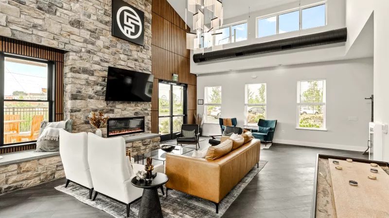Stylish resident clubhouse at The Foundry featuring a double-height ceiling, stone fireplace with mounted TV, cozy seating areas, modern chandelier, and a shuffleboard table for community enjoyment.