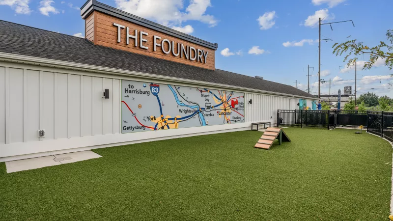 Fenced artificial turf dog park with agility ramp, seating area, and colorful Lancaster-area map mural under a clear blue sky.