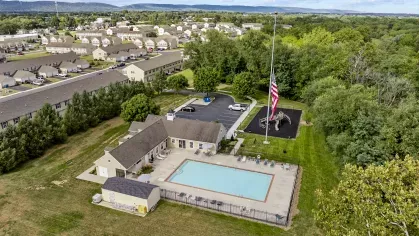 An aerial drone view of the clubhouse and swimming pool at Keystone Arms in Carlisle, PA. The photo shows the resort-style pool with a large sun deck, lounge chairs, and the community clubhouse building. In the background, there is a parking lot and a playground, all set against a backdrop of lush green trees.