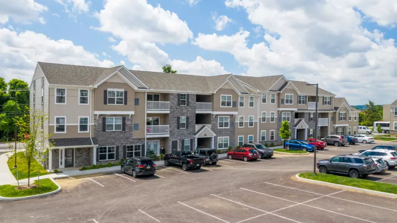 Three-story apartment building with beige and gray siding, multiple balconies, and parked cars in a spacious parking lot under a partly cloudy sky.