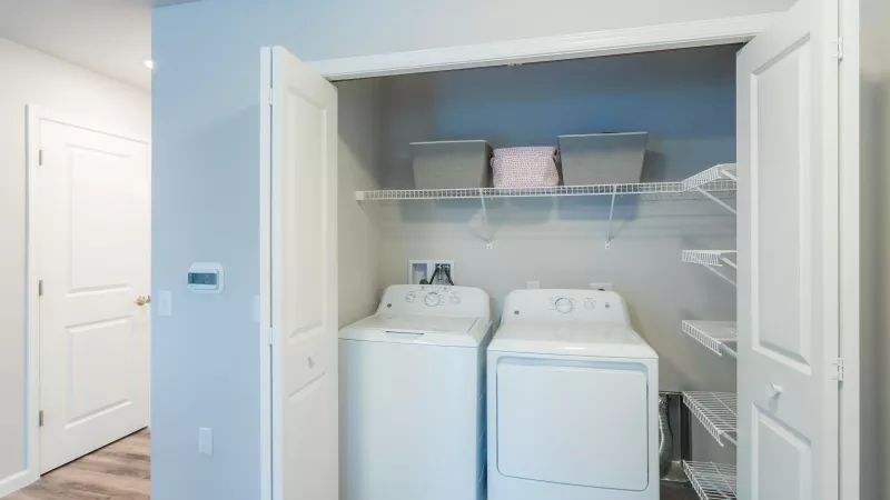 In-home laundry at Wyndham Pointe Apartments showing full-size washer and dryer with shelving and folding storage space inside a white double-door closet.