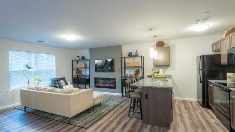 Open-concept living area at Wyndham Pointe Apartments featuring a cozy sectional sofa, modern electric fireplace with mounted TV, and contemporary kitchen with granite island and pendant lighting.