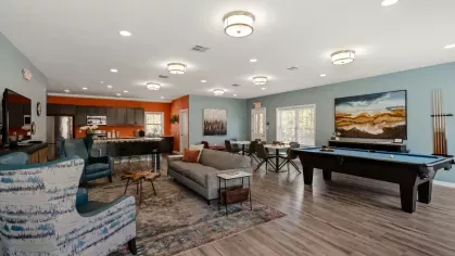 Clubhouse lounge area with modern furniture, pool table, dining tables, and wood-look flooring.