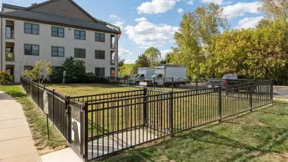 Fenced outdoor dog park area with grass and gated entry next to the apartment building and surrounding trees.