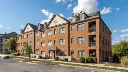 Three-story brick apartment building with dormer windows, fenced patios, and landscaped walkways under a blue sky.