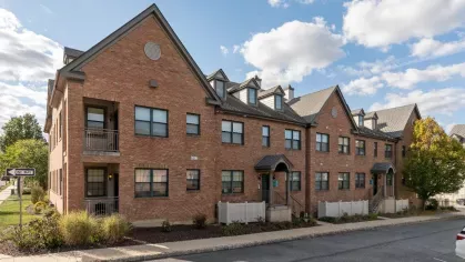 Row of brick townhome-style apartments with covered entrances, balconies, and a sidewalk along the street.