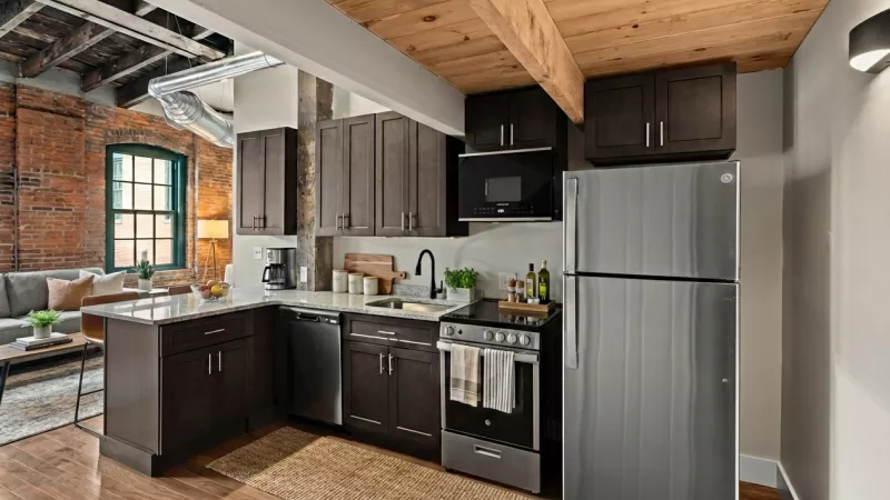 A modern kitchen with dark wood cabinetry, granite countertops, and stainless steel GE appliances. The design highlights industrial elements like exposed ductwork and rustic wooden ceiling beams.