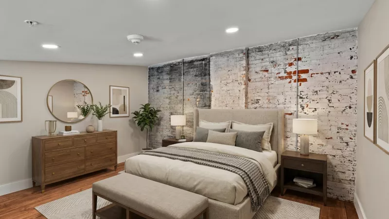 A bedroom featuring an authentic whitewashed brick accent wall and warm wood flooring. The room is illuminated by recessed lighting and offers a neutral color palette.