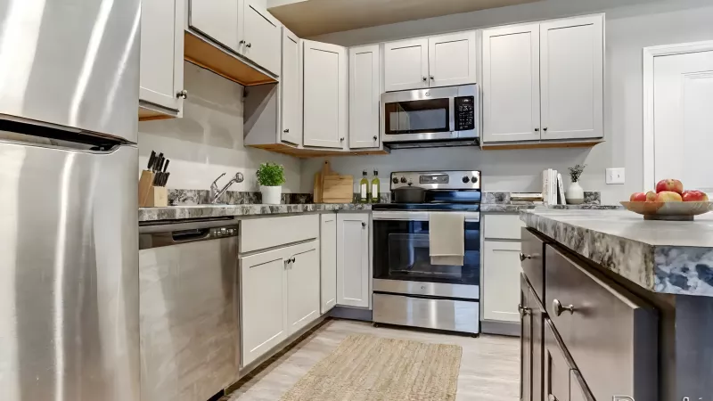 A bright kitchen with stainless steel appliances, including a refrigerator, dishwasher, and stove. The light grey cabinetry is accented by dark grey granite-style countertops and a neutral-toned backsplash. A tan woven rug is centered on the light-colored flooring between the cabinets and a dark wood kitchen island.