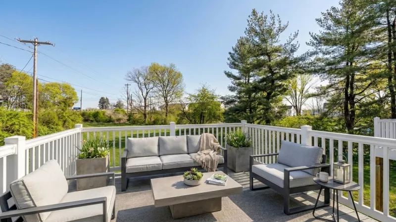An elevated wooden deck with white railings overlooking a green lawn and mature trees. The deck is furnished with a grey outdoor sofa and matching armchairs.