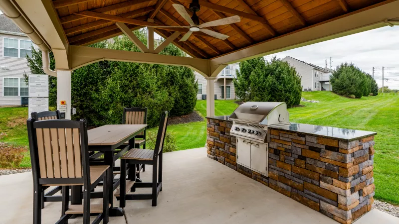 A covered outdoor grilling station featuring a built-in stainless steel gas grill set into a stacked-stone base with dark countertops. The area is protected by a large wooden pavilion with a ceiling fan and includes a high-top dining table with four chairs.