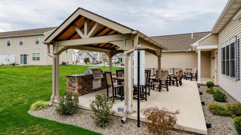 A wide-angle view of a concrete community patio featuring a covered grilling area and several rows of Adirondack-style lounge chairs. The space is situated behind residential buildings with light-colored siding and overlooks a shared green space.