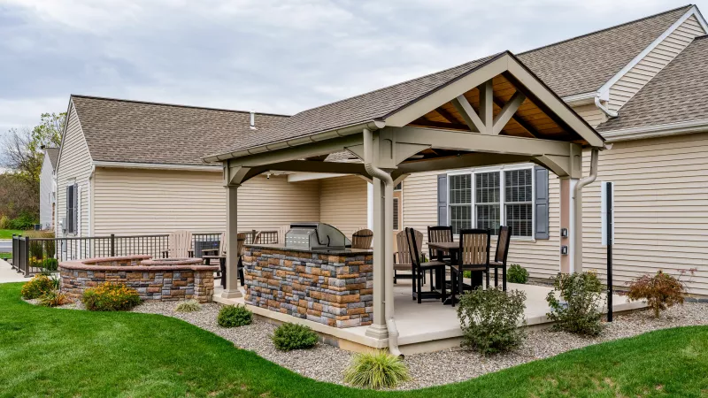 An outdoor amenity space featuring a large wooden pavilion and a circular stacked-stone fire pit. The area is surrounded by manicured green lawns and professional landscaping with gravel borders.