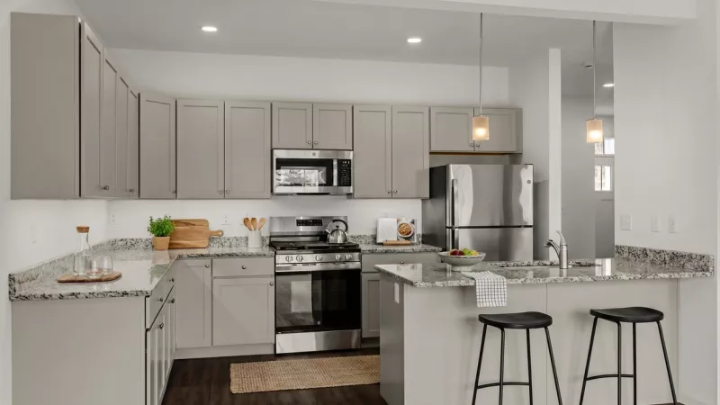 A close-up of a contemporary kitchen showcasing light grey cabinets, speckled granite countertops, and a stainless steel gas range and microwave. An island with a breakfast bar and modern black stools provides additional seating.
