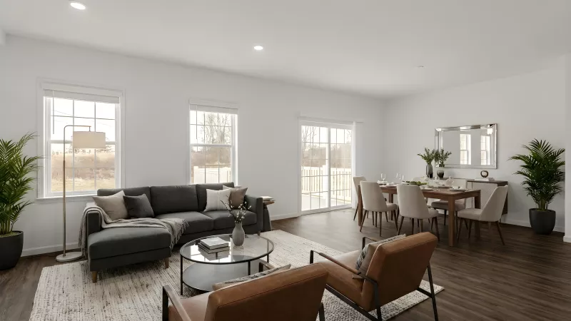 An expansive living and dining area with dark wood-style flooring and white walls. The room is illuminated by natural light from large windows and features a modern dining set and a comfortable seating area.