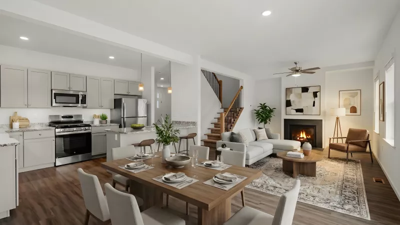 A wide-angle view of a modern open-concept living area featuring a kitchen with light grey cabinetry, granite countertops, and stainless steel appliances. The space flows into a dining area and a living room with a fireplace and large windows.