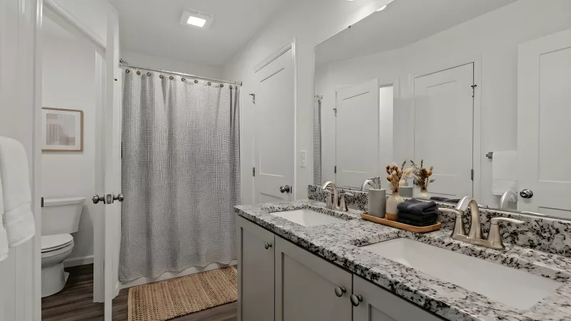 A primary bathroom featuring a double vanity with a granite countertop and brushed nickel faucets. The room includes a large mirror, a separate toilet area, and a shower with a textured neutral curtain.