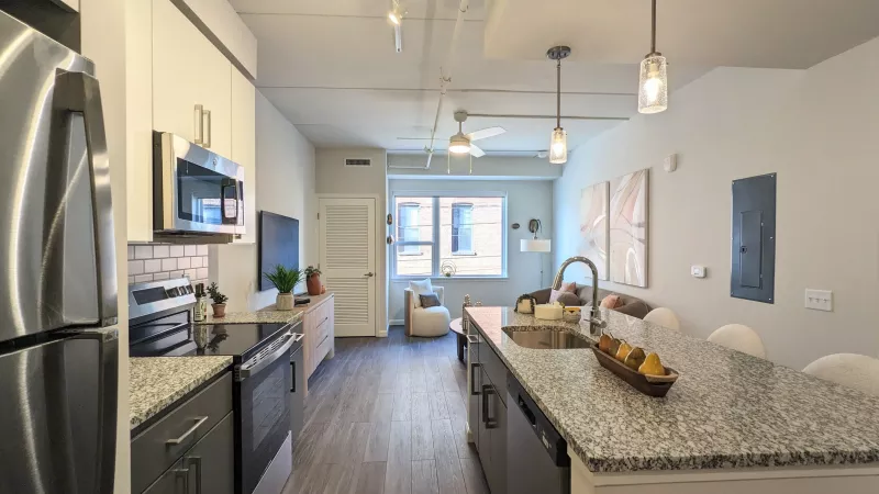 An open-concept layout showing a kitchen with a large granite-style island, stainless steel appliances, and gray lower cabinetry transitioning into the living area.