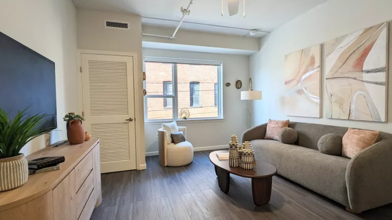 A modern living room featuring dark wood-style flooring, a large window, and industrial-style exposed ceiling pipes. The space includes a neutral sofa and a light-wood media console.