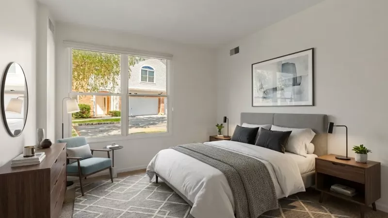 A bright, modern bedroom featuring a large window that overlooks the community, showing a neighboring townhome with an attached white garage door. The room is styled with neutral tones, a gray upholstered bed frame, and wooden nightstands.