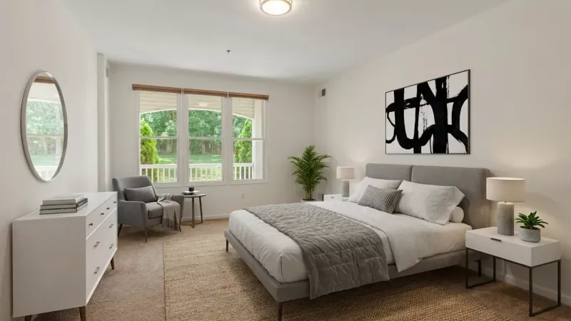 A sunlit primary bedroom featuring a neutral color palette and a large three-pane window with a view of a private outdoor balcony and mature green trees. The room is furnished with a gray bed, a white dresser, and modern minimalist decor.