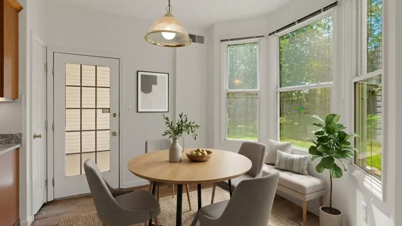 A cozy dining nook featuring a large bay window that provides a bright view of the surrounding greenery. The space includes a round wooden table with four chairs, a modern hanging light fixture, and a glass-paneled door leading outside.