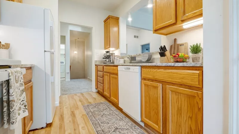 A view of a kitchen with light oak cabinets, white dishwasher, and white refrigerator. A pass-through window looks into an adjacent living area, and the space is finished with light wood-style flooring and a neutral-patterned runner rug.