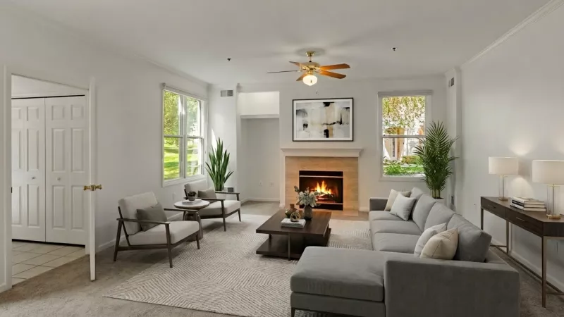 A spacious living room with neutral-toned carpeting, white walls, and a central fireplace with a tan stone surround and white mantel. The room includes a ceiling fan with integrated lighting and two large windows that provide a view of the exterior landscaping.