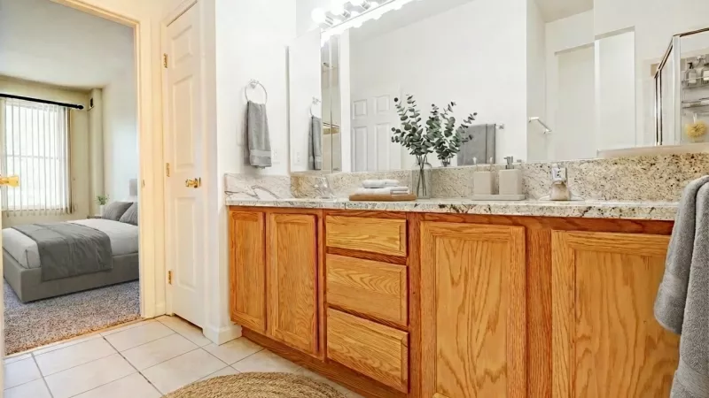 A large bathroom featuring a long oak vanity with a granite-style countertop and double sinks. A wide mirror with theater-style lighting spans the wall above the vanity. The flooring is white square tile, and the room includes a view into an adjacent carpeted bedroom with a window.