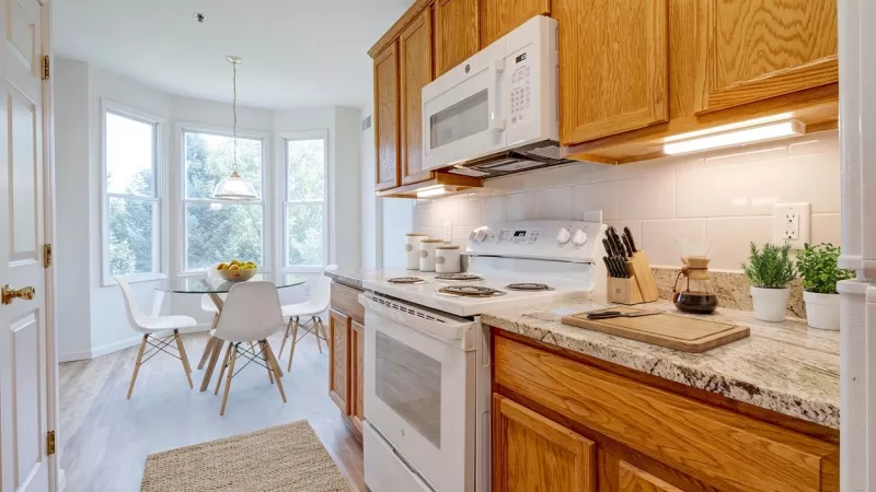 A galley-style kitchen featuring oak-finish cabinetry, granite-patterned countertops, and white appliances including an electric range and over-the-range microwave. The kitchen opens into a bright dining nook with a bay window overlooking green trees, featuring light wood-style plank flooring.