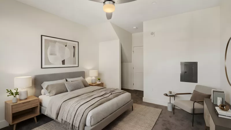 A cozy bedroom with neutral plush carpeting and off-white walls. The room features a gray upholstered bed frame, light wood nightstands, a ceiling fan, and a dedicated sitting nook with a neutral armchair and a small side table.