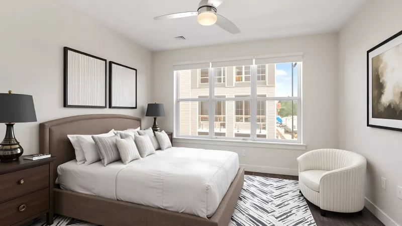 A spacious bedroom featuring a large window that overlooks the apartment complex exterior. The room includes a dark wood bed frame with white bedding, matching nightstands with black lamps, a ceiling fan, and a modern patterned area rug over dark flooring.