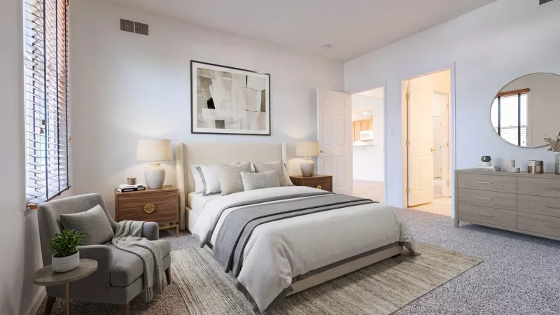 A well-lit primary bedroom featuring neutral-colored walls and carpet, a large window with blinds, a queen-sized bed with plush bedding, and a separate sitting area with an armchair.