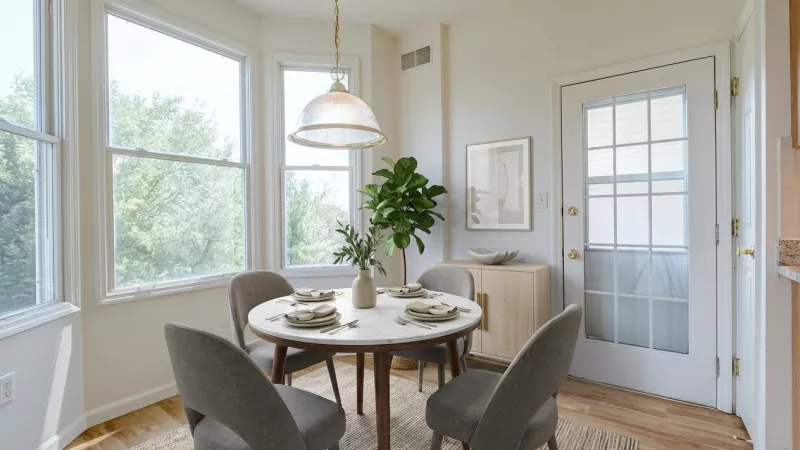 A bright dining nook with a circular table and four chairs, featuring three large windows that provide a view of the surrounding greenery.