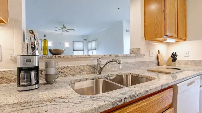 A modern kitchen featuring granite-style countertops, classic wood cabinetry, a double stainless steel sink, and an open breakfast bar overlooking the living area.