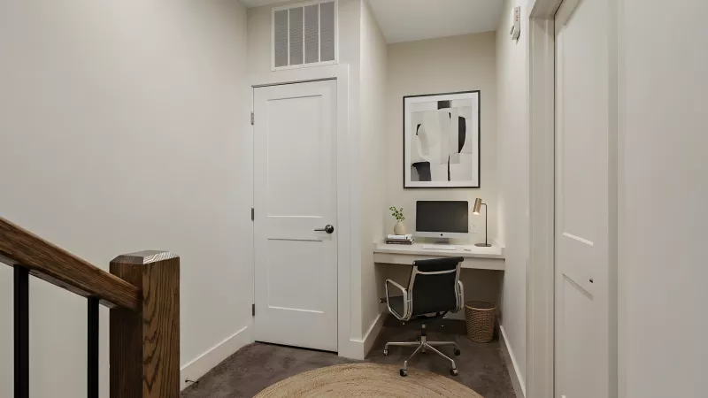 A functional built-in desk nook located in a carpeted hallway area. The space features a white countertop desk, a modern black office chair, overhead recessed lighting, and a neutral color palette.