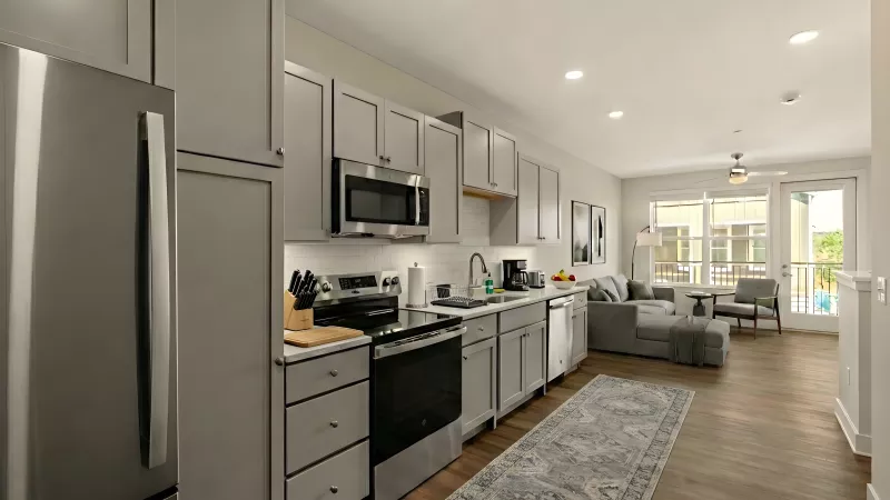 A wide-angle view of an open-concept living area featuring light wood-style flooring. The kitchen includes gray cabinetry, stainless steel appliances, and a white subway tile backsplash. The space flows into a living room with a gray sectional sofa and a glass door leading to a private balcony.