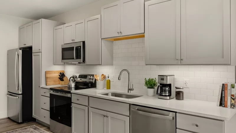 A close-up of a modern kitchen with gray shaker-style cabinets and white quartz countertops. It features a stainless steel dishwasher, an oven with a glass cooktop, a built-in microwave, and a white subway tile backsplash with under-cabinet lighting.