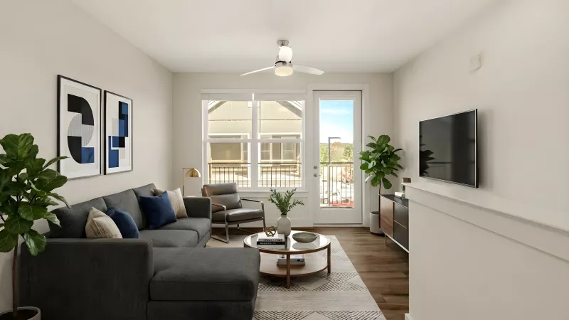 A bright living room with neutral walls and a large window and glass door providing natural light. The room is styled with a dark gray sectional, a round wooden coffee table, and a wall-mounted TV above a decorative ledge.