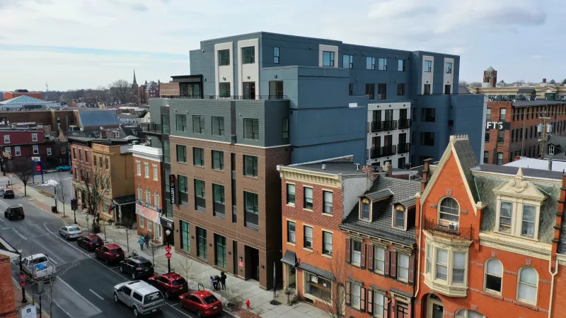Elevated view of a modern blue multi-story building alongside older brick rowhouses and parked cars on a city street.