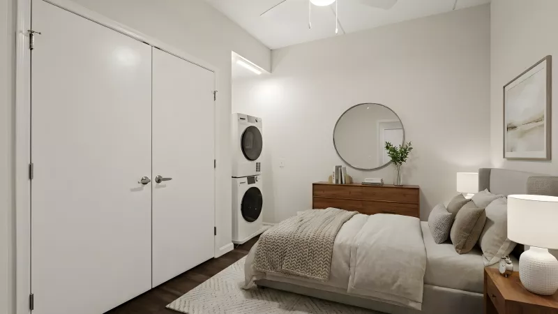 Modern bedroom with a bed, wood dresser, round mirror, and a stacked washer/dryer. Dark wood floor, light walls, and ceiling fan.