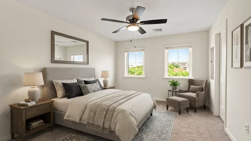Bright bedroom with a gray upholstered bed, wooden nightstand, armchair, two windows, and a ceiling fan.