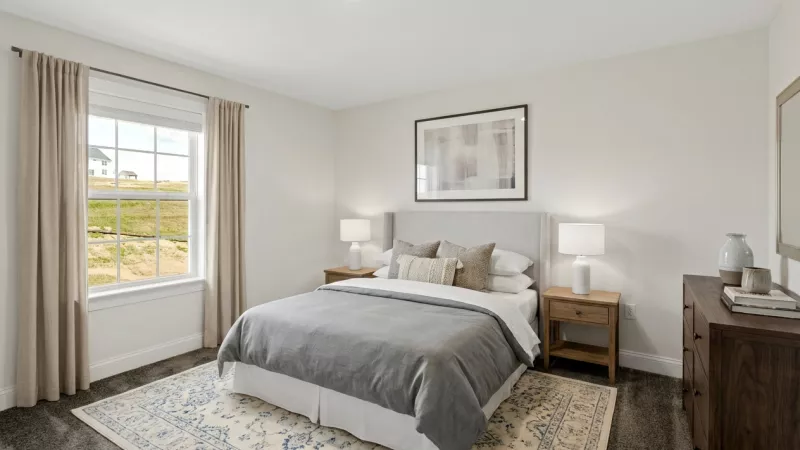 Well-lit bedroom with a grey-duveted bed, two nightstands with lamps, a large window, and a blue and beige patterned rug.