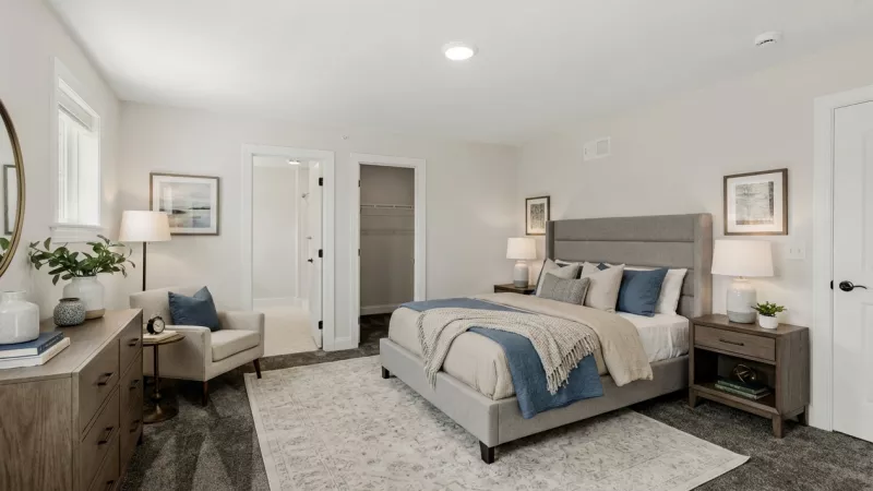 Modern bedroom with a grey upholstered bed, blue accents, dresser, armchair, and light walls. Doors open to other rooms.