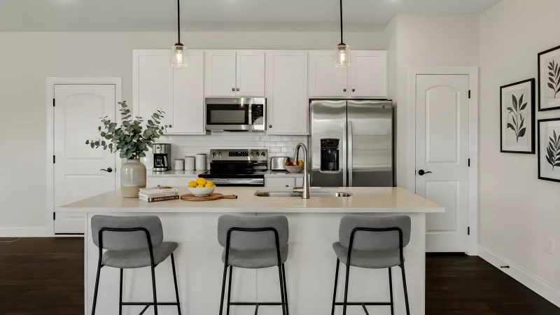 Modern white kitchen with island, gray stools, stainless steel appliances, and dark wood floor.