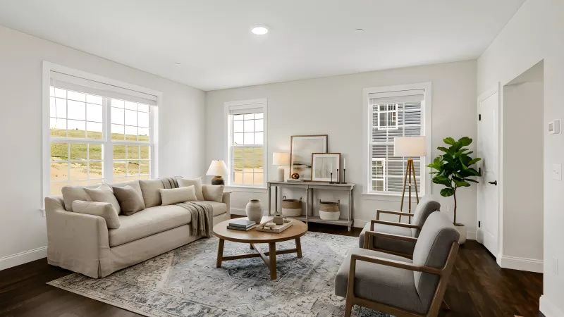 Bright living room with a beige sofa, round coffee table, two grey armchairs, console table, and a blue patterned rug on dark wood floors.