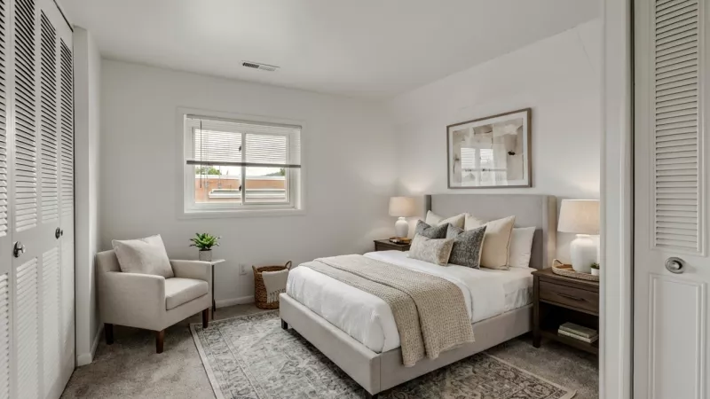 Bright bedroom with a bed, two nightstands, lamps, a window, and an armchair. White closet doors frame the room.