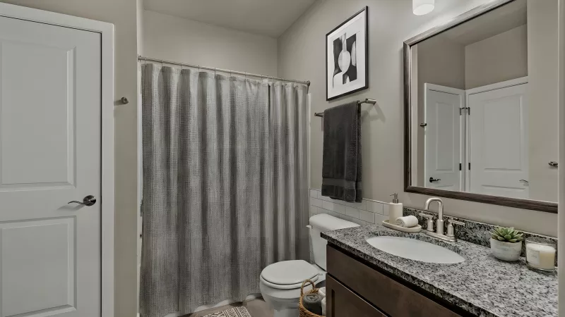 Modern bathroom with a dark wood vanity, speckled counter, white sink, mirror, toilet, and gray shower curtain.