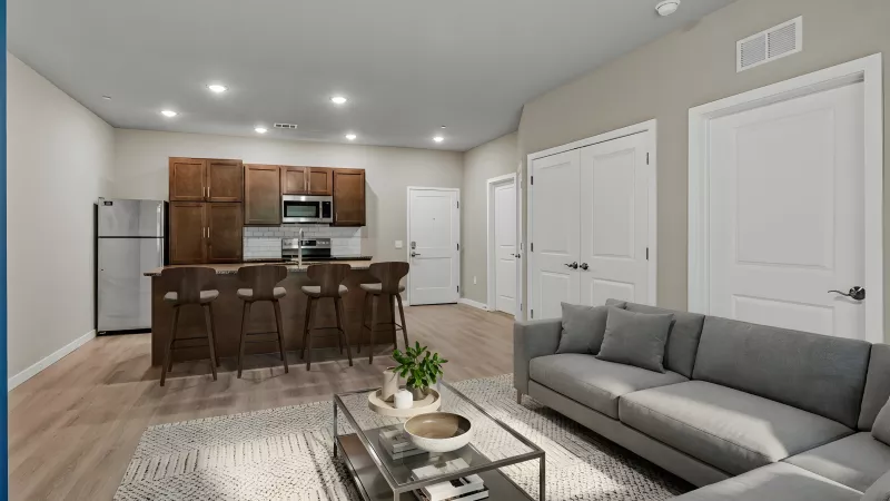Modern open-plan living room with grey sectional sofa, glass coffee table, and a kitchen featuring dark wood cabinets and a breakfast bar.