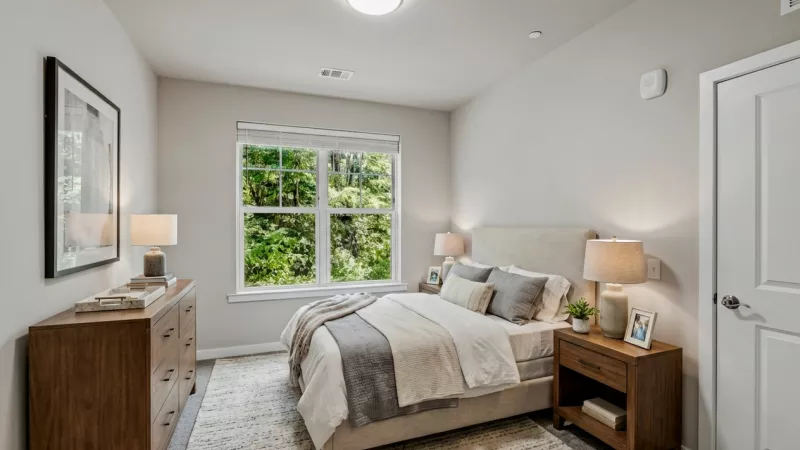 Modern bedroom with a neutral queen bed, wood furniture, and a bright window looking out to green foliage.